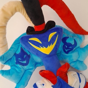 Op de afbeelding: Een pluche speelgoed met een blauw personage met een gele lachende gezicht, een zwarte hoge hoed en rode en witte accenten. Het personage heeft lichtblauwe vleugels met een donkerblauw embleem. Het speelgoed is gemaakt van zachte stof.