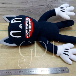 May include: A plush toy in the shape of a cartoon cat with a large, toothy grin. The cat is blue with white gloves and a gray face. The toy is lying on a wooden surface with a ruler in the foreground. The ruler has numbers 1 through 30 printed on it.