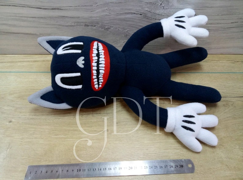 May include: A plush toy in the shape of a cartoon cat with a large, toothy grin. The cat is blue with white gloves and a gray face. The toy is lying on a wooden surface with a ruler in the foreground. The ruler has numbers 1 through 30 printed on it.