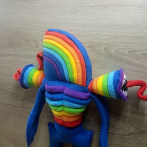 Puede incluir: Un juguete de peluche de color arco&iacute;ris con un cuerpo largo y delgado y una cabeza grande. El juguete tiene una cabeza de color arco&iacute;ris y un torso de color arco&iacute;ris. El juguete tiene una cola larga y delgada y dos brazos largos y delgados. El juguete est&aacute; hecho de tela suave y de felpa.