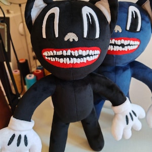 Pode incluir: Um brinquedo de pelúcia de gato preto com olhos brancos grandes, uma boca vermelha larga com muitos dentes brancos e mãos com luvas brancas. Um segundo brinquedo de pelúcia semelhante em azul escuro está posicionado atrás da figura preta.