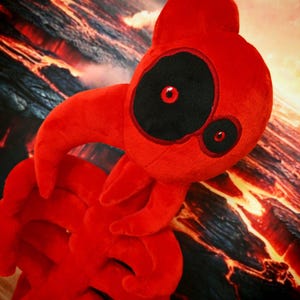 May include: A plush red creature with a large black eye and smaller eye, both with red pupils. The creature has a bulbous head and multiple appendages. The background features a fiery, lava-like landscape.