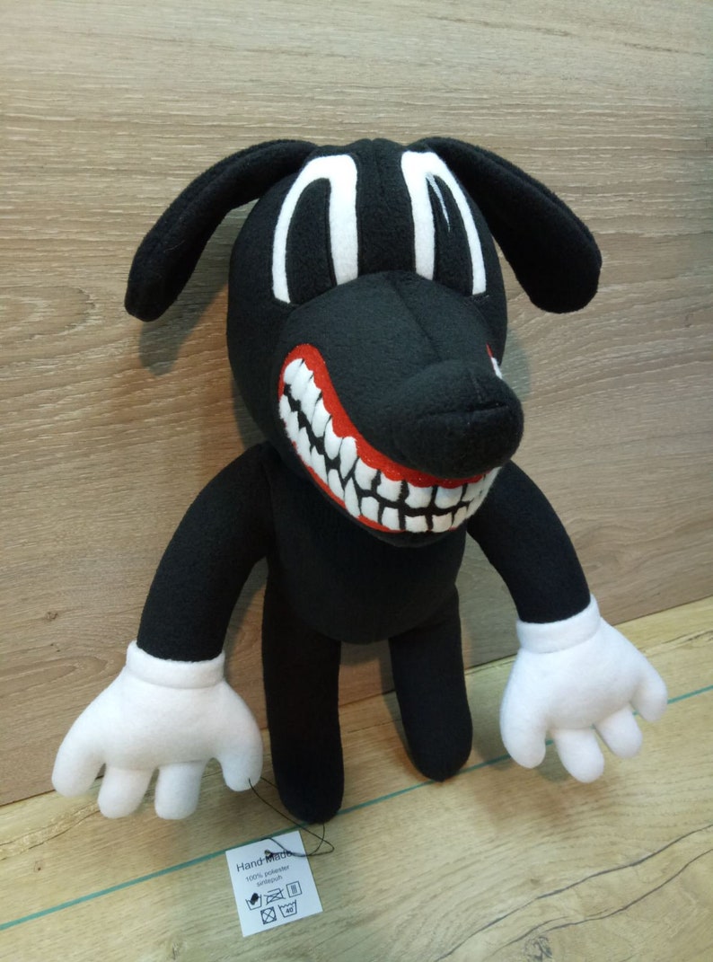 May include: Black plush cartoon dog with a large red mouth and white teeth. The dog has white eyes and white gloves. The dog is standing on a wooden surface. A tag attached to the dog reads 'Hand-Made 100% polyester  cotton' with icons for washing instructions.