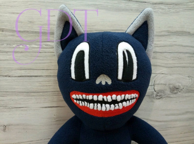 May include: A plush toy cat with a blue body, gray ears, and a large white smile with red lips. The cat's eyes are large and black with white outlines.