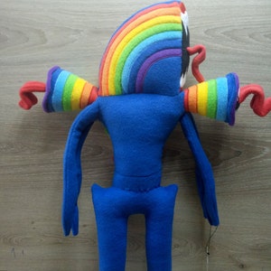 Puede incluir: Un juguete de peluche azul con brazos de arco&iacute;ris y una cabeza de arco&iacute;ris. El juguete tiene brazos y piernas largos.