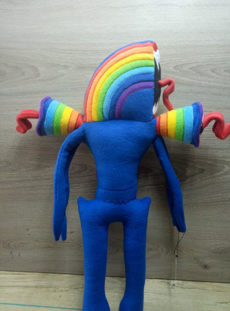Puede incluir: Un juguete de peluche azul con brazos de arco&iacute;ris y una cabeza de arco&iacute;ris. El juguete tiene brazos y piernas largos.