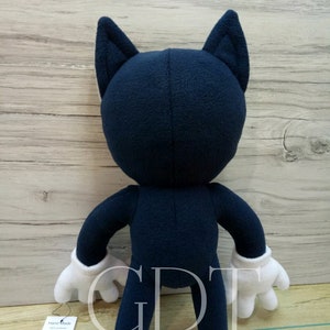 May include: A plush toy in the shape of a cartoon character. The toy is blue with white gloves and large ears. The toy is standing on a wooden surface. The toy has a tag that says 'Hand Made'.