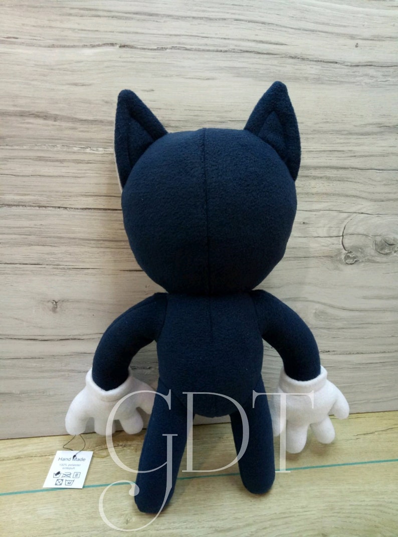 May include: A plush toy in the shape of a cartoon character. The toy is blue with white gloves and large ears. The toy is standing on a wooden surface. The toy has a tag that says 'Hand Made'.