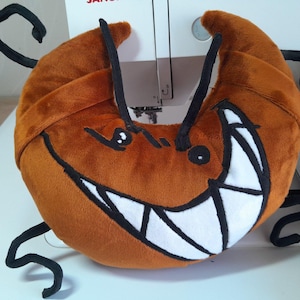 May include: A plush, orange, crescent-shaped creature with a wide, toothy grin and black, curved antennae. The toy has black eyes and is likely handmade. The background includes a sewing machine and spools of thread.