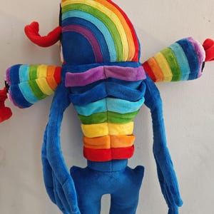 Pode incluir: Um brinquedo de pelúcia com corpo azul e torso segmentado exibindo listras horizontais nas cores do arco-íris. A criatura tem dois braços largos com mangas listradas de arco-íris, apêndices vermelhos nas laterais e uma cabeça curva.