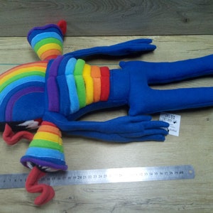 Puede incluir: Juguete de peluche arco&iacute;ris con extremidades azules y un torso arco&iacute;ris. El juguete est&aacute; acostado sobre una superficie de madera con una regla para la escala. El juguete mide aproximadamente 76 cm de largo.