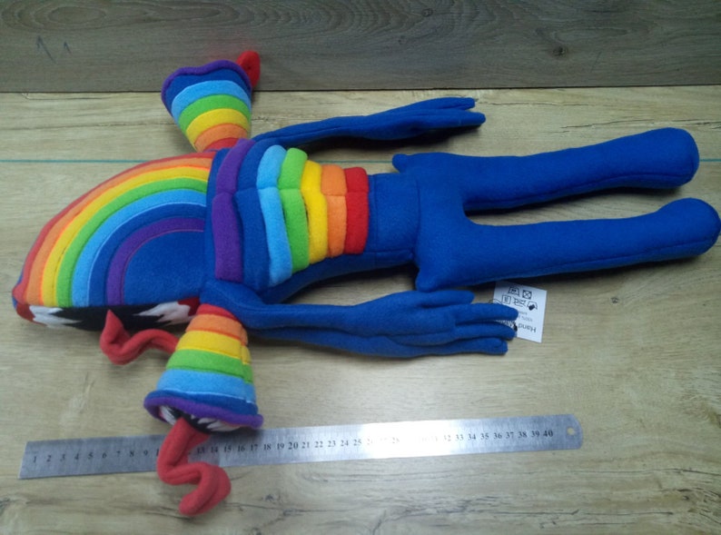 Puede incluir: Juguete de peluche arco&iacute;ris con extremidades azules y un torso arco&iacute;ris. El juguete est&aacute; acostado sobre una superficie de madera con una regla para la escala. El juguete mide aproximadamente 76 cm de largo.