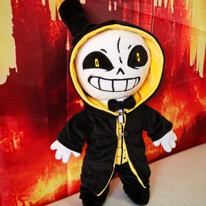 Puede incluir: Un peluche de un personaje con una cara blanca con forma de calavera, ojos negros y una amplia sonrisa. El personaje lleva un sombrero de copa negro, un abrigo negro con ribetes amarillos y una capucha forrada de amarillo. El fondo es rojo y naranja.