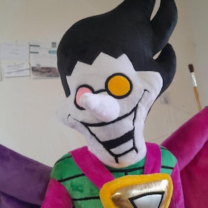 May include: A plush toy of a character with green and pink clothing, a yellow and gold triangle on the chest, and a white face with a large, toothy grin and one yellow eye.