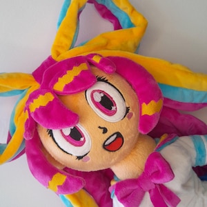 Könnte beinhalten: Plüschpuppe mit einem Cartoon-Gesicht, großen rosa Augen und rosa Haaren mit gelben Akzenten. Die Puppe trägt ein weißes und blaues Outfit mit einer rosa Schleife. Das Haar ist eine Mischung aus Rosa, Gelb und Blau.