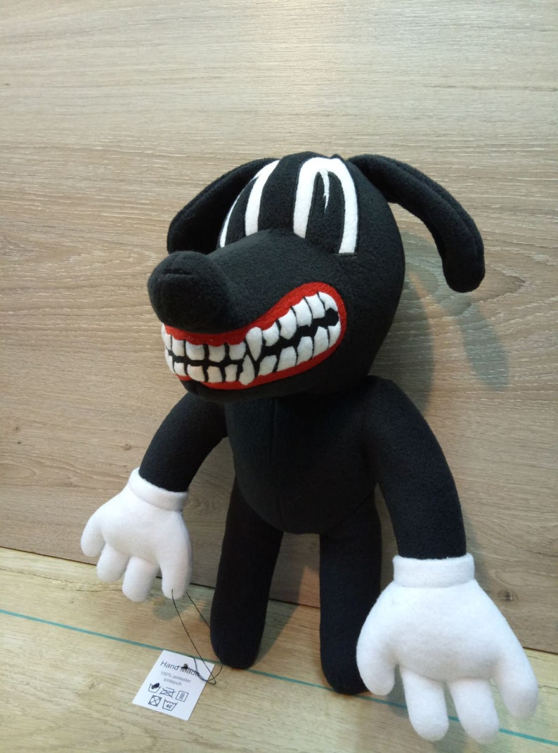 May include: A black plush cartoon dog with white eyes, a red mouth with white teeth, and white gloves. The dog is standing on a wood surface. The tag attached to the dog reads 'Handmade with love, wash delicate, do not bleach, do not tumble dry, iron low, do not dry clean.'
