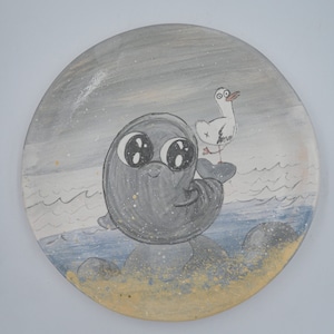 May include: A hand-painted ceramic plate with a gray seal and a white seagull on a blue and yellow beach scene.
