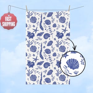 May include: A white beach towel with a repeating pattern of blue seashells, starfish, and seaweed. The towel is hanging on a clothesline against a blue sky background. The towel has a close-up view of a seashell with the text "FUCK TRUMP".
