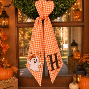 May include: A long, orange and white gingham fabric ribbon hanging from a green wreath. The left tail features an embroidered white ghost with an orange bow, while the right tail displays a large, black embroidered letter H with flourishes.