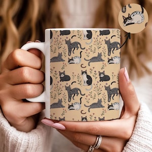 May include: White ceramic mug featuring a repeating pattern of various cat illustrations in gray, black, and white on a tan background. The cats are depicted in different poses, with floral accents. The mug is held by a person.
