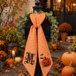 May include: A black mailbox adorned with a green eucalyptus wreath and a long orange and white gingham ribbon. The ribbon features an embroidered black letter M on one side and a jack-o'-lantern with bats and a crescent moon on the other.