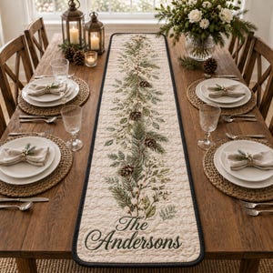 May include: A long, rectangular, quilted table runner with a cream-colored background and a central botanical print featuring green pine branches, white berries, and brown pinecones. The text "The Andersons" is printed in a dark script font.
