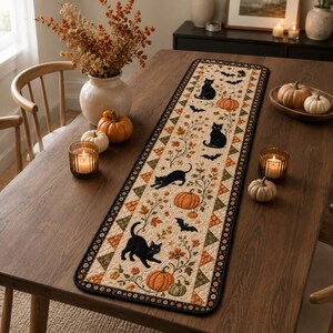May include: A rectangular quilted table runner featuring a cream-colored background with a repeating pattern of black cats, orange pumpkins, and black bats. The border consists of small triangles in shades of orange, green, and cream.