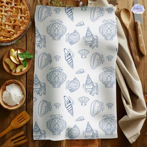 May include: White kitchen towel with a blue seashell pattern. The towel features various seashell illustrations, including conch shells and scallop shells. The words "Fred & George" are printed on the towel. The towel is on a wooden surface.