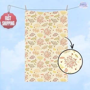 May include: A cream-colored towel with a repeating pattern of autumn leaves in shades of brown, yellow, and green. Some leaves have the text "FUCK TRUMP" inside. The towel is hanging on a clothesline against a blue sky.