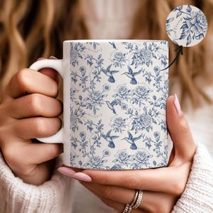 May include: White ceramic mug featuring a blue floral and hummingbird pattern. The design includes detailed illustrations of flowers, hummingbirds, and delicate vines. The mug has a standard handle and a smooth, glossy finish.