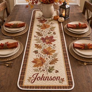 May include: A rectangular quilted table runner featuring a central print of autumn leaves, acorns, and the name Johnson in script. The runner has a plaid border and is set on a wooden table with four place settings, each with a plate and napkin.