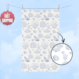 May include: A white beach towel with a repeating pattern of blue seashells, coral, and small circles. The towel is hanging on a clothesline. A red and white tag with the words "FAST SHIPPING" is attached to the line.