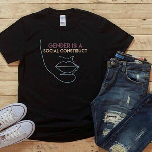 May include: Black t-shirt with the text "GENDER IS A SOCIAL CONSTRUCT" and a line drawing of a face. The shirt is paired with blue jeans and white sneakers on a wooden surface.