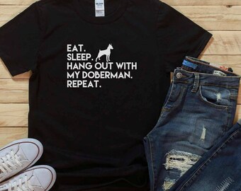 Op de afbeelding: Zwart T-shirt met witte tekst die luidt "Eten. Slapen. Hang out met mijn Dobermann. Herhalen."  Een wit silhouet van een Dobermann bevindt zich boven de tekst.