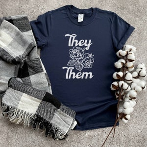 Pode incluir: Uma camiseta azul marinho com um design floral branco e o texto "They Them" impresso na frente.