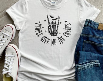 Puede incluir: Camiseta blanca con un gráfico negro de una mano de esqueleto haciendo un signo de rock and roll. El gráfico está rodeado de estrellas y murciélagos y el texto "People Give Me The Creeps".
