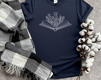 Puede incluir: Una camiseta azul marino con un contorno blanco de un libro abierto con flores que salen de él.