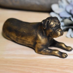以下が含まれることがあります： 横たわっている犬の金色の金属製置物。犬は短いしわくちゃの顔と短い尻尾をしています。
