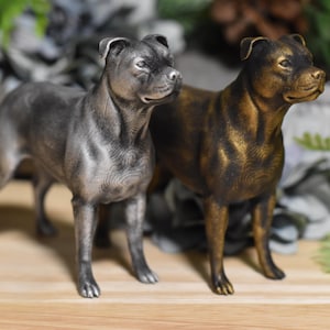 May include: Two silver and gold painted Staffordshire Bull Terrier dog figurines. The figurines are standing on a wooden surface.