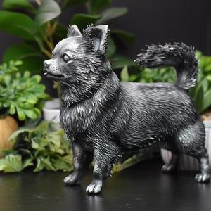 Puede incluir: Una figurita de un chihuahua de color plateado. La figurita est&aacute; de pie sobre una superficie negra y mira hacia la izquierda. El perro tiene una cola larga y tupida y est&aacute; de pie con las patas delanteras ligeramente separadas.