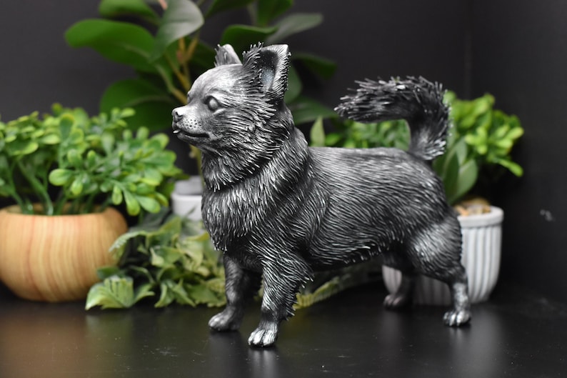 Puede incluir: Una figurita de un chihuahua de color plateado. La figurita est&aacute; de pie sobre una superficie negra y mira hacia la izquierda. El perro tiene una cola larga y tupida y est&aacute; de pie con las patas delanteras ligeramente separadas.