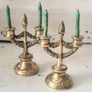 May include: Two miniature brass candelabras featuring a central finial and two candle holders with textured green candles topped with red wicks. The metal bases and arms are decorated with embossed laurel leaf patterns and a weathered finish.