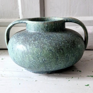 May include: A round ceramic vase featuring two arched handles on opposite sides. The exterior surface has a mottled, speckled finish in shades of forest green, sage, and hints of blue, creating a textured appearance across the entire vessel body.