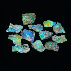 Pode incluir: Uma coleção de pedras preciosas de opala bruta, exibindo flashes vibrantes de cor. As opalas exibem uma mistura de azuis, verdes e amarelos, em um fundo escuro. Cada pedra tem uma textura natural única.
