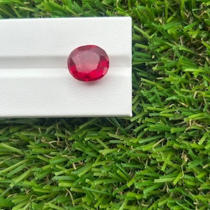 May include: A vibrant, oval-cut red gemstone with a faceted surface that reflects light, displayed on a white rectangular stand. The gemstone exhibits a deep, saturated crimson hue and a polished finish, showcasing its clarity and internal structure.