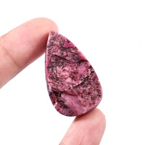 Peut inclure: Une pierre précieuse polie en forme de goutte d'eau, avec un mélange de teintes roses et brun foncé. La surface de la pierre présente un motif marbré, avec des variations de couleur et de texture. La pierre est tenue entre deux doigts sur fond blanc.