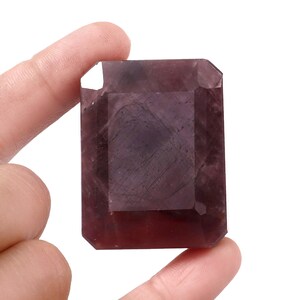 Puede incluir: Una piedra preciosa rectangular facetada con un tono púrpura oscuro y marrón rojizo. La piedra presenta una superficie pulida con inclusiones internas visibles y un diseño de bordes planos biselados, mostrando una calidad translúcida.