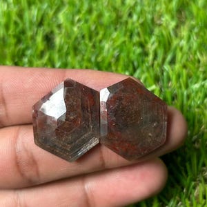Puede incluir: Dos piedras preciosas hexagonales facetadas de un tono marrón rojizo profundo con inclusiones internas visibles. Las piedras presentan una superficie superior plana con múltiples facetas angulares en los bordes, mostrando un aspecto semitranslúcido.