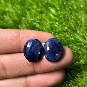Könnte beinhalten: Zwei oval geformte, tiefblaue Saphire mit facettierter Oberfläche. Jeder Edelstein zeigt eine dunkle, gesättigte Farbe mit sichtbarer innerer Textur und lichtreflektierenden Facetten auf der Oberseite, präsentiert als ein Paar loser Steine.
