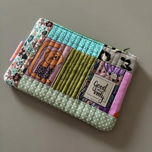Könnte beinhalten: Eine grüne, rosa und blaue Patchwork-Zipper-Tasche mit einer Katzenillustration und einem weißen Etikett mit der Aufschrift "Good Vibes Only".